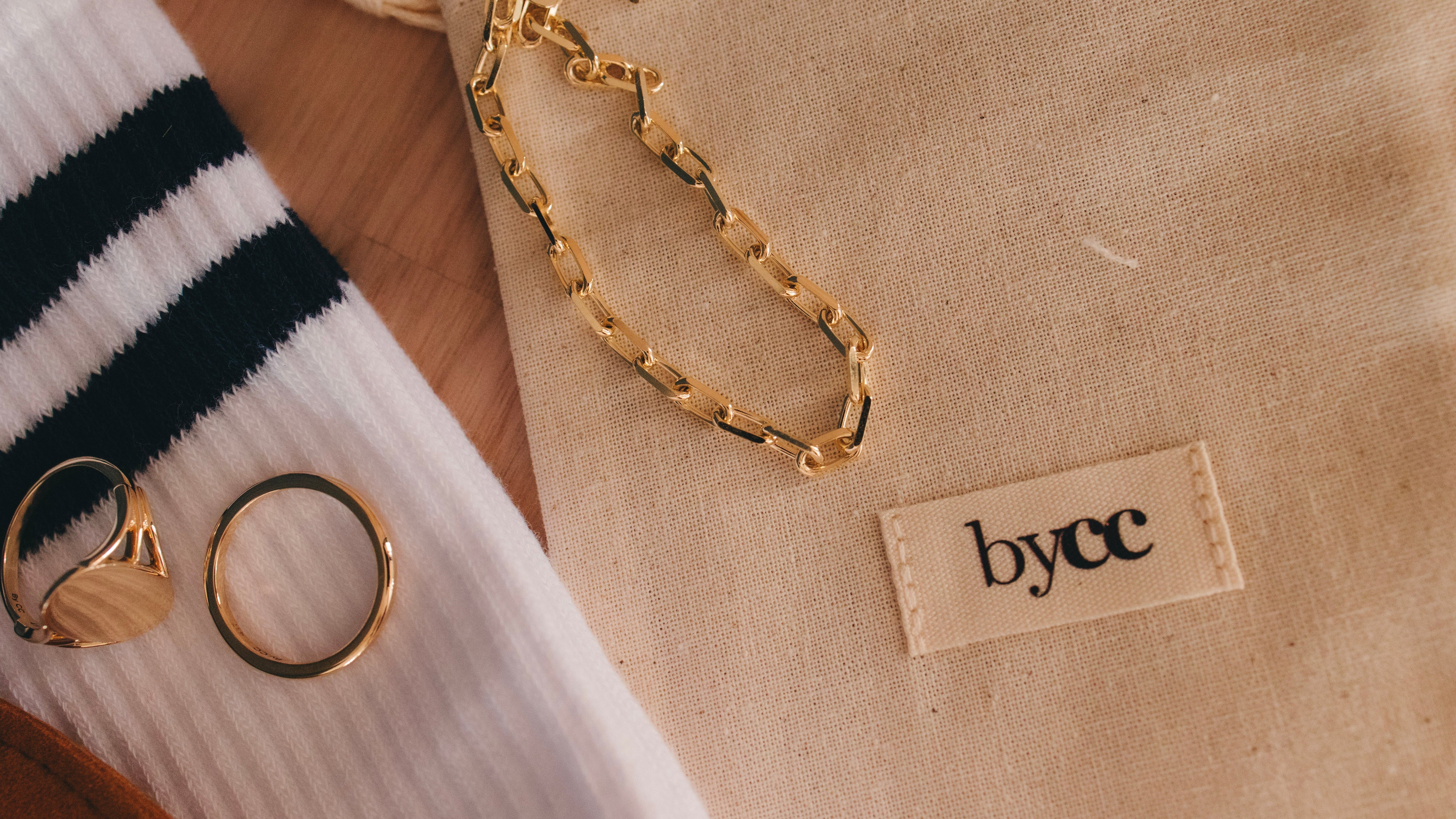 Gold chain and rings on a beige fabric with 'bycc' label