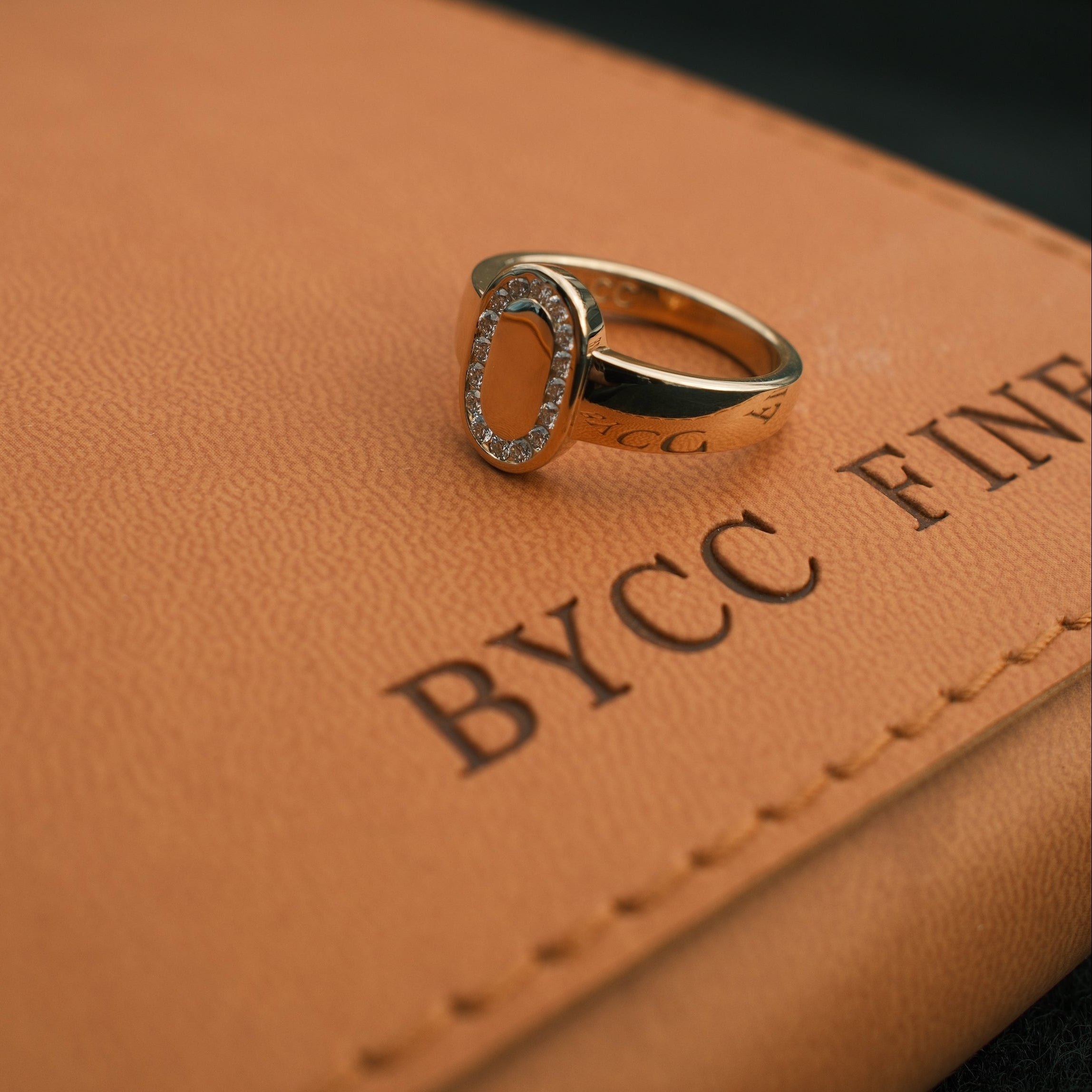 Close-up of a gold and diamond ring on a brown leather surface with 'ByCC' branding.