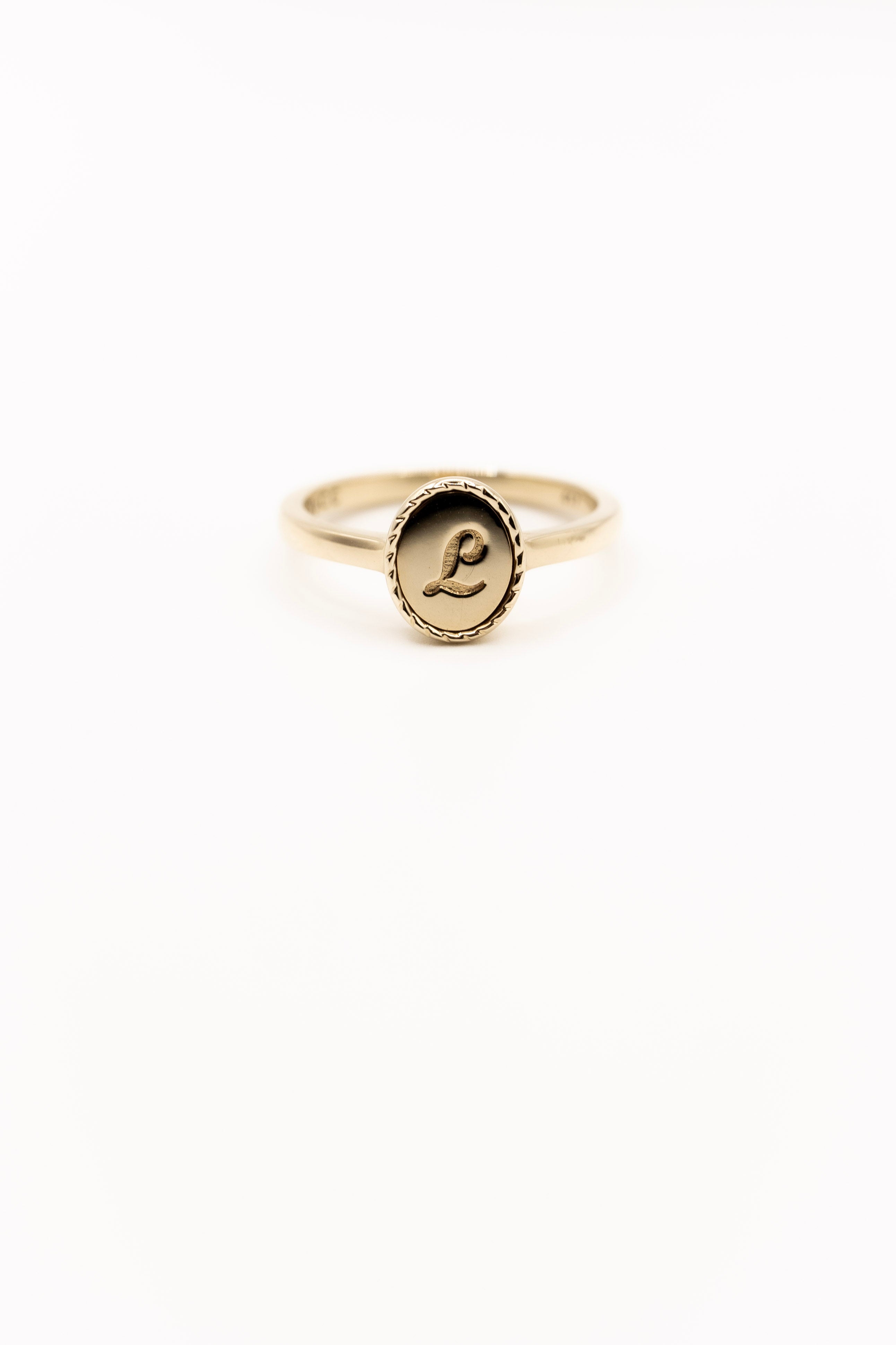 Gold ring with a circular emblem featuring a letter 'L' on a white background
